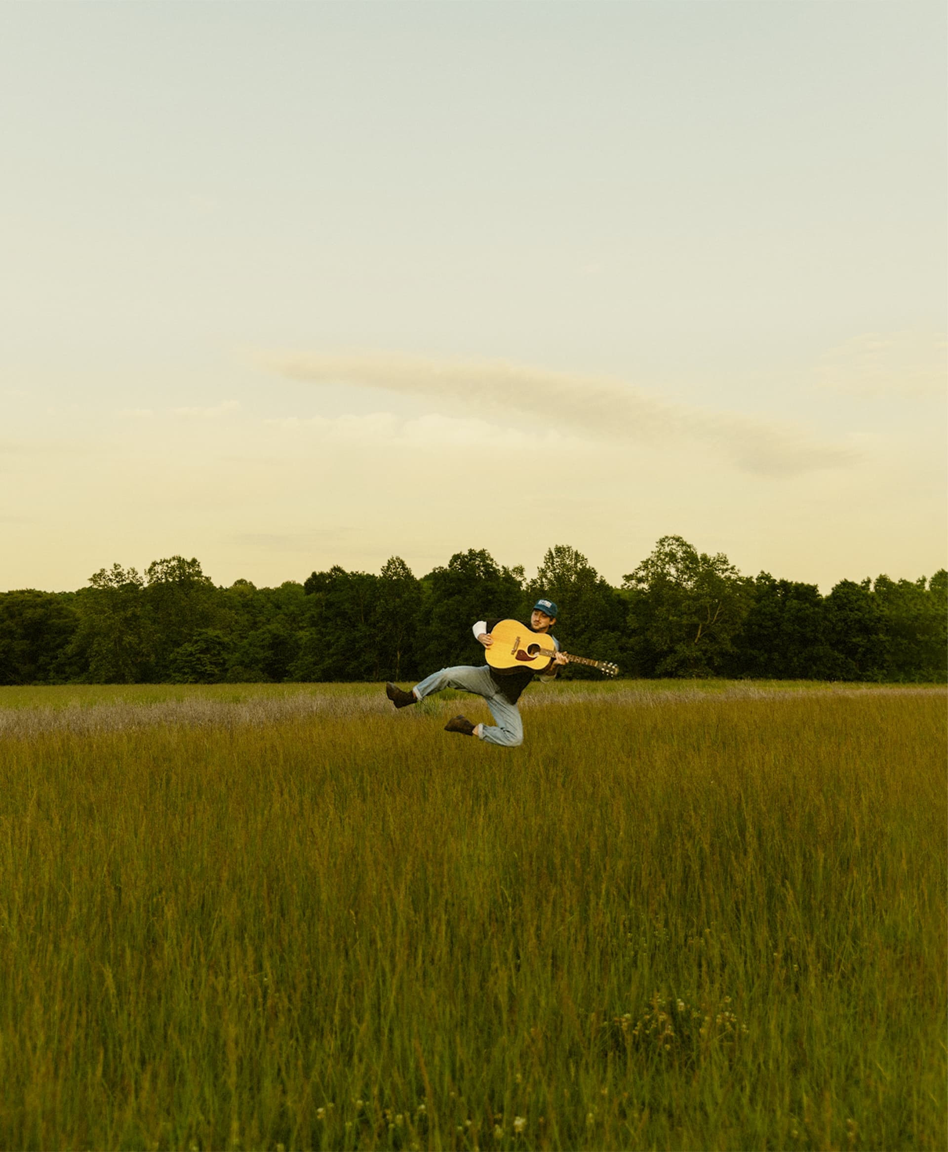 Hunter Flynn mid-jump with his guitar in an open field at dusk